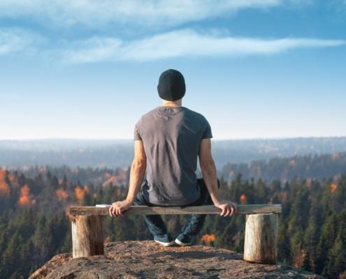 Mann sitzt auf einer Bank und genießt die atemberaubende Aussicht auf einen herbstlichen Wald, während er ein Einweg Vape verwendet. Das Bild symbolisiert Ruhe, Entspannung und den Genuss moderner Entspannungstechniken in der Natur.