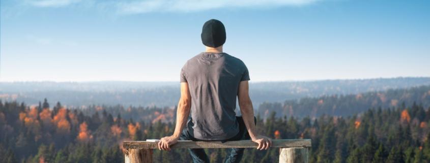 Mann sitzt auf einer Bank und genießt die atemberaubende Aussicht auf einen herbstlichen Wald, während er ein Einweg Vape verwendet. Das Bild symbolisiert Ruhe, Entspannung und den Genuss moderner Entspannungstechniken in der Natur.