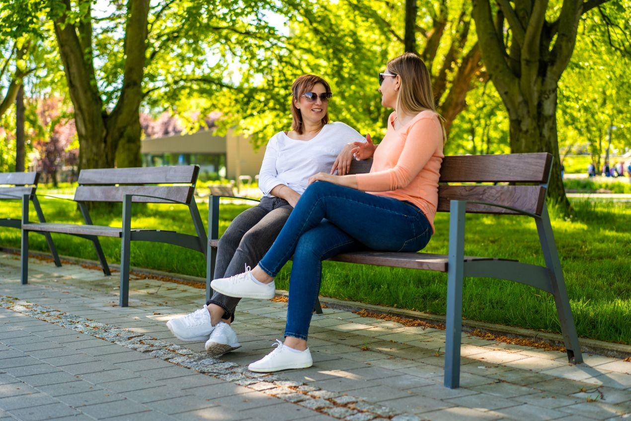 Zwei Frauen sitzen auf einer modernen Parkbank in einem sonnigen Park und unterhalten sich entspannt im Grünen.