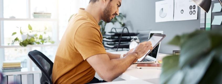 Mann arbeitet im Buero mit Tablet an Content-Planung, Erklaervideo als Teil der Landingpage-Strategie sichtbar im Hintergrund