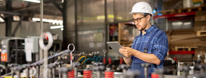Mitarbeiter mit Helm nutzt Produktionsplanung Software auf Tablet in moderner Fertigungshalle